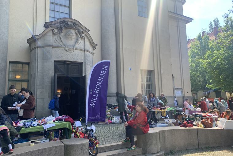 Flohmarkt vor dem Portal der Hoffnungskirche mit Tischen voller Bücher und Kinderkleidung und einer lila Willkommen-Fahne